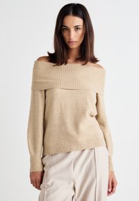 Beige off-shoulder sweater with ribbed neckline and cuffs. Soft texture and relaxed fit, paired with light-colored pleated trousers.