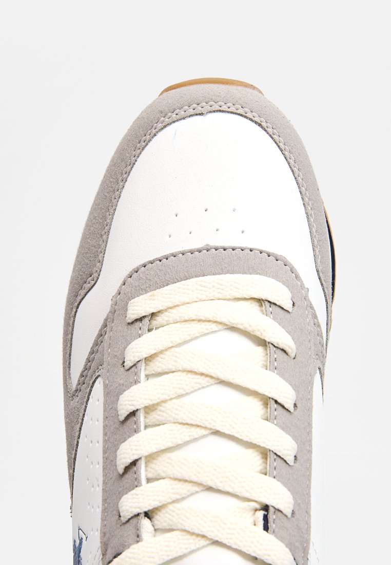 Zapatilla que presenta una parte superior de cuero blanco predominantemente, con acentos de gamuza gris, cordones planos color beige y una suela de goma marrón texturizada.