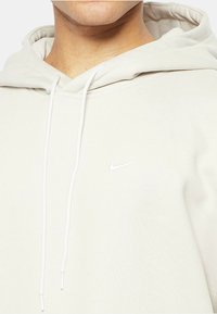 Ljust beige huva sweatshirt tillverkad av mjukt tyg, med en framficka och en vit Nike-logotyp på bröstet. Dragsko kopplad till huvan.