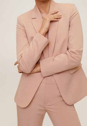 Osoba ubrana w jasno różowy, dopasowany żakiet i pasujące spodnie, z jedną ręką założoną na ciało, a drugą spoczywającą blisko kołnierza.