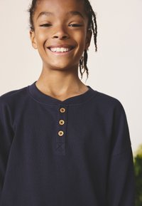 Navy blue ribbed shirt featuring a round neckline, three wooden buttons, and loose sleeves. Textured fabric with a soft finish.