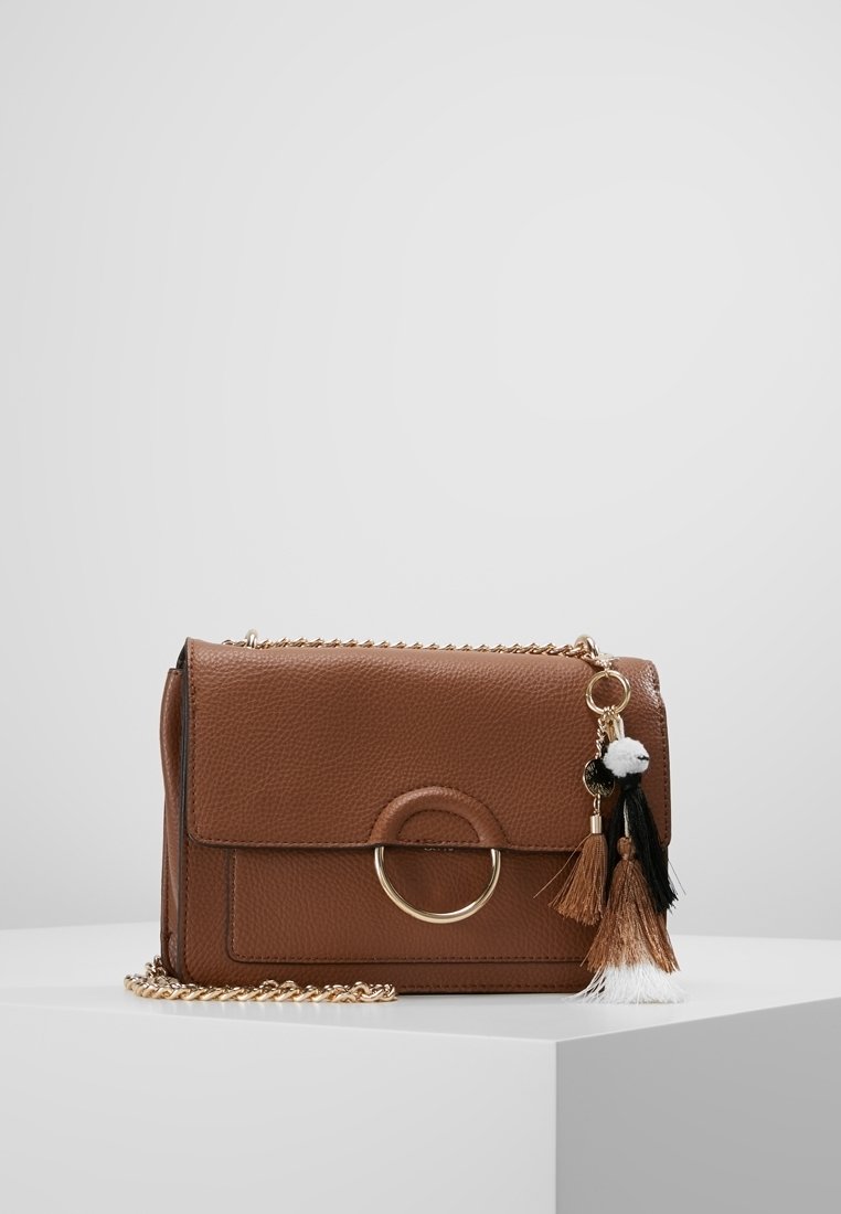 Borsa a tracolla in pelle marrone con chiusura a patta, accentata da un anello dorato e nappine decorative in nero, bianco e marrone. Inclusa tracolla a catena.