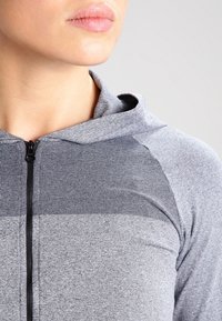 Femme portant un sweat à capuche zippé gris avec un panneau côtelé sur la poitrine et une capuche ouverte, sur un fond gris clair.