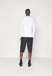 Weißes Langarmshirt und dunkelgraue, kurze Sportshorts. Die Shorts haben einen umgeschlagenen Saum und werden durch schwarze Socken und Sportschuhe ergänzt.
