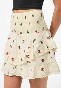 Jupe courte crème avec des couches en volants, présentant un motif floral cerise et un tissu texturé. Taille élastique avec ourlet festonné.