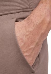 Braune Sporthose mit seitlicher Tasche und gerippten Details sowie einer glatten Stofftextur, die die Hand in der Tasche hervorhebt.