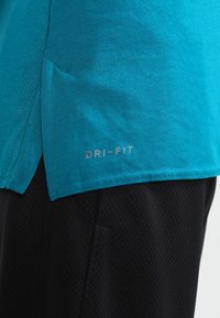 Primo piano dell'orlo di una maglietta sportiva teal con il testo "DRI-FIT" sopra a pantaloni corti neri texturizzati.