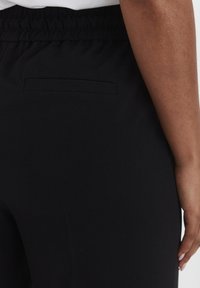 Zwarte broek heeft een elastische tailleband en een platte, welvingen zak achterin. De stof is glad en heeft een lichte glans.