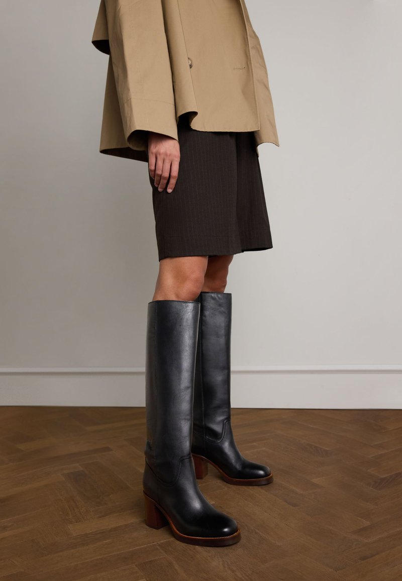 Tall black leather boots with a smooth finish, square toe design, and a chunky wooden heel. Worn with a brown pinstripe skirt and beige jacket.