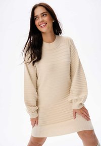Gebreide crèmekleurige jurk met ribbelstructuur, ronde halslijn en volumineuze mouwen. Eindigt boven de knie met een subtiele band aan de zoom.