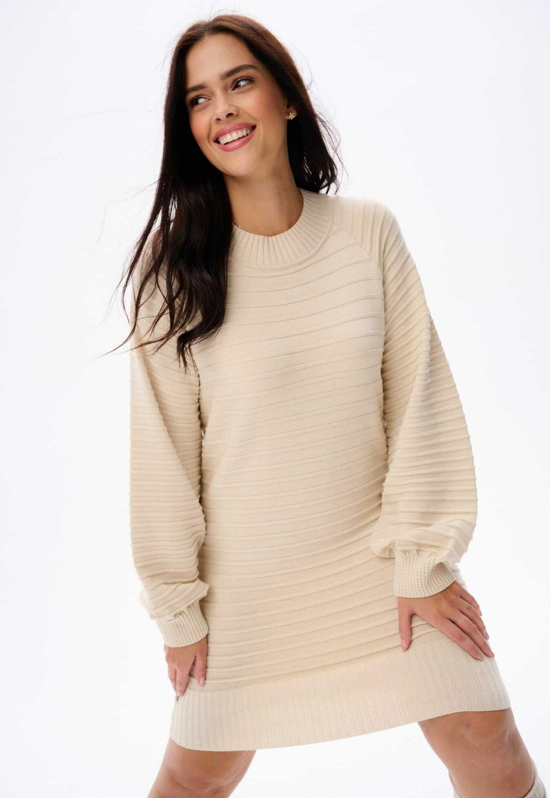 Gebreide crèmekleurige jurk met ribbelstructuur, ronde halslijn en volumineuze mouwen. Eindigt boven de knie met een subtiele band aan de zoom.