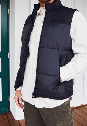 Mężczyzna noszący czarną pikowaną kamizelkę z logo "HILFIGER", białą sztruksową koszulę i oliwkowe spodnie typu cargo, stojący w pomieszczeniu obok zielonych drzwi.