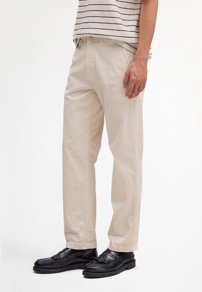 Person trägt beige Straight-Leg-Hosen, schwarze Ledermokassins und ein weißes Hemd mit dünnen schwarzen horizontalen Streifen.