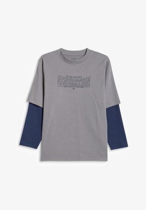 Chłopiec w zielonej czapce typu beanie, szarej oversize koszulce z napisem "Generation", granatowych długich rękawach i spodniach oraz białych trampkach, stojący na szarym tle.