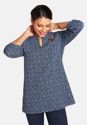 Frau in blau gemusterter Tunika mit gesticktem Schmetterling richtet lächelnd sanft ihr Haar vor schlichtem weißem Hintergrund.