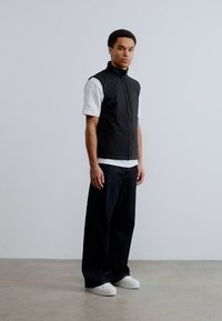 Άνδρας που στέκεται μέσα σε κλειστό χώρο, φορώντας μαύρο αμάνικο γιλέκο πάνω από λευκό μπλουζάκι, φαρδιά μαύρη παντελόνια και λευκά αθλητικά παπούτσια, μπροστά σε μια απλή ανοιχτόχρωμη τοίχο.