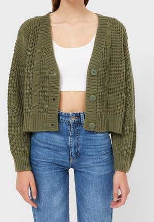 Femme portant un cardigan court en maille épaisse vert olive avec de gros boutons, par-dessus un débardeur blanc court et un jean taille haute bleu.