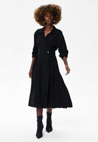 Kobieta z kręconymi włosami, ubrana w długą czarną suknię z rękawami do łokci i czarnymi botkami za kostkę, stojąca na białym tle.