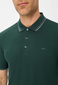 Dunkelgrünes Polohemd aus strukturiertem Stoff, mit einer Knopfleiste mit drei Knöpfen, einem kontrastierenden weißen Kragen und einem "Brax"-Logo auf der Brust.