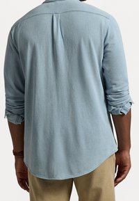 Ljusblå texturerad skjorta med långa ärmar, uppskjutna manschetter med en knappdetalj och rundad nederkant. Bakre veckdesign ger en subtil detalj.