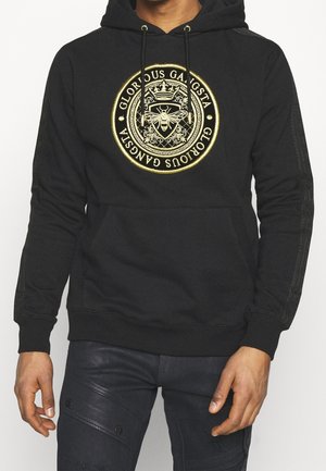 Person trägt einen schwarzen Hoodie mit einem goldenen, kreisförmigen Emblem, das eine Biene, eine Krone und den Schriftzug "Glorious Gangsta" auf der Vorderseite zeigt.