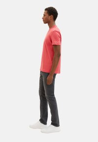 Rotes Baumwoll-T-Shirt mit Rundhalsausschnitt, kurzen Ärmeln und einem Logo-Detail am linken Ärmel, kombiniert mit dunklen Jeans und weißen Turnschuhen.