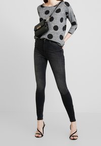 Harmaa pitkähihoinen paita, jossa on suuria mustia pilkkuja, yhdistettynä tummiin slim fit -farkkuihin ja mustiin nilkkureihin. Pienikokoinen musta olkalaukku on käytössä.