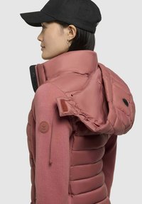Gevoerde roze jas met een afneembare capuchon en elastische accenten, voorzien van een zwarte rits en logopatch op de mouw. Fleece textuur op de mouwen.