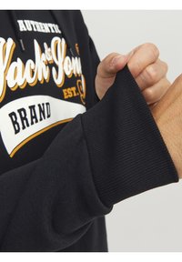 Felpa nera con un logo "Jack & Jones" bianco e arancione. Il polsino è a coste, enfatizzando la texture del capo. Primo piano della manica.