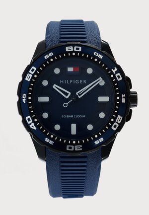 Montre analogique bleue Tommy Hilfiger avec cadran rond, index blancs, lunette tournante et bracelet en silicone texturé bleu.