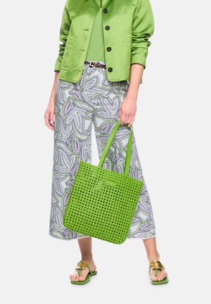 Woman wearing green jacket and patterned wide-leg pants, holding woven green tote bag, and wearing green sandals with gold buckle.
