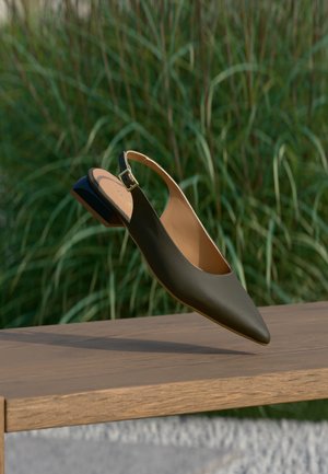 Μαύρο δερμάτινο παπούτσι τύπου slingback με μυτερή μύτη και μικρό τακούνι, αιωρούμενο πάνω από ξύλινο παγκάκι σε φόντο πράσινου χόρτου.