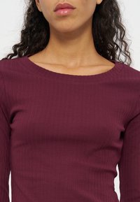 Femme portant un haut violet foncé côtelé à manches longues avec un large col arrondi, laissant apparaître une partie de son cou et de sa clavicule.