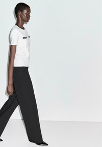 Woman walking in black wide-leg trousers, white short-sleeve top with black trim, and black pointed shoes against a plain white background.