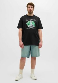 Camiseta de algodón negra con un estampado gráfico de una estatua rodeada de follaje verde. Combinada con pantalones cortos de mezclilla azul claro y zapatillas blancas.