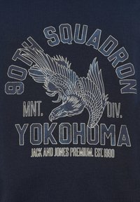 Μπλε ναυτικό μπλουζάκι με γραφικό αετού με λεπτομερή φτερά, περιτριγυρισμένο από το κείμενο "90TH SQUADRON MNT. DIV. YOKOHOMA." σε λευκό.