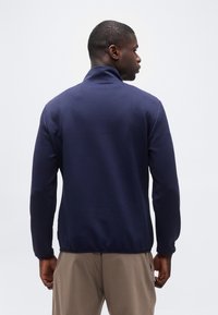 Άνδρας με σκούρο μπλε σακάκι και μπεζ παντελόνι, στέκεται γυρισμένος με την πλάτη σε απλό λευκό φόντο.