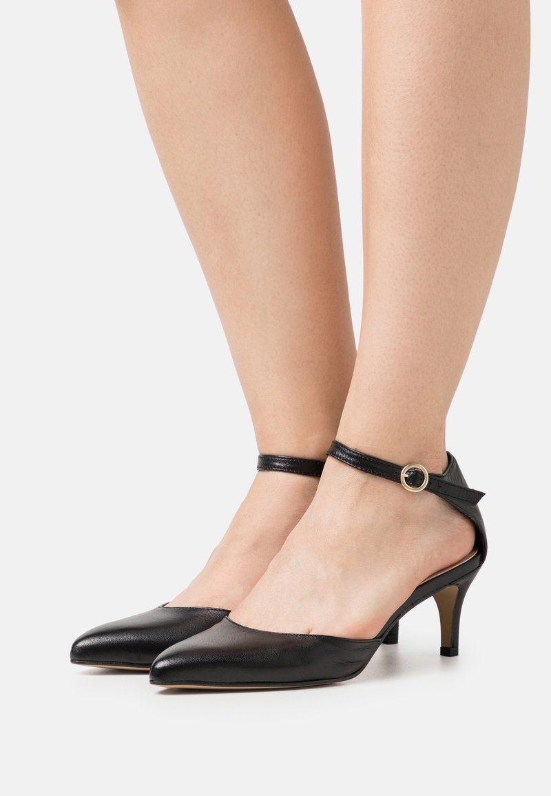 Pieds portant des talons hauts noirs à bout pointu avec des brides à la cheville et de petites boucles dorées, présentés sur un fond clair uni.