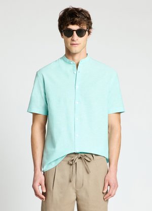 Mann trägt schwarze runde Sonnenbrille, hellblaues kurzärmeliges Hemd mit Mandarinkragen und beige Hose mit Kordelzug vor weißem Hintergrund.