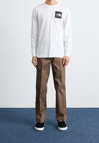 Bílá košile s dlouhým rukávem a černým logem na hrudi, v kombinaci s hnědými kalhotami. Černé boty doplňují outfit. Jednoduché pozadí.