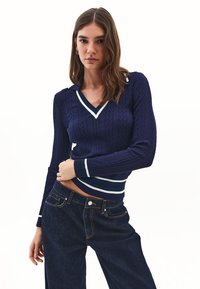 Suéter de punto azul marino con diseño de trenzas, cuello en V y cuello y puños a rayas verde y blanco; combinado con jeans de mezclilla oscuros de cintura alta.