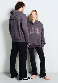 Dwóch młodych dorosłych w szarych bluzach z kapturem i czarnych spodniach, stojących obok siebie na tle jednolitej białej ściany.