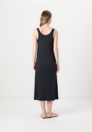 Mujer de pie, de espaldas, vestida con un vestido midi negro sin mangas y zapatos planos beige, sobre un fondo claro y uniforme.