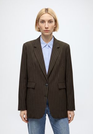 Femme aux cheveux blonds courts portant un blazer marron à fines rayures sur une chemise bleu clair à col et un jean bleu, debout devant un fond uni.