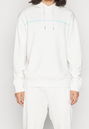 Osoba ubrana w białą bluzę z kapturem i spodnie dresowe z jasnoniebieskim paskiem oraz małym napisem na piersi i "AX" na spodniach.