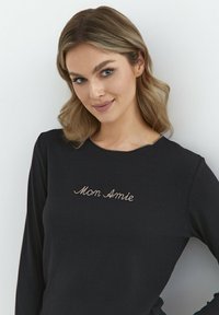 Czarna koszula z długim rękawem z miękkiego materiału, z złotym haftowanym napisem "Mon Amie" na piersi, z okrągłym dekoltem.
