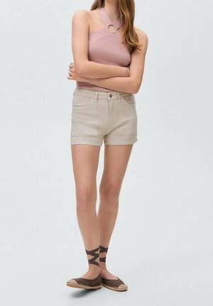 Woman wearing a pink halter top with ring detail, beige high-waist shorts, and brown espadrilles with ankle ribbons, standing with arms crossed.