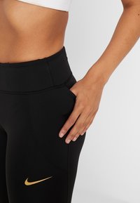 Leggings neri a vita alta con una texture liscia e elasticizzata. Presentano il logo dorato Nike sulla gamba sinistra e dettagli con tasche laterali.