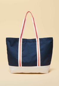 Tumesinine ja valge kandekott, millel on pikad valged sangad ja punased triibud, seisab beezil pinnal püstiasendis.