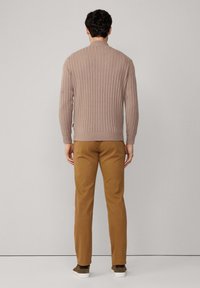 Beżowy sweter z prążkowanej tkaniny z wysokim kołnierzem, zestawiony z jasnobrązowymi spodniami o zwężanym kroju i ciemnobrązowym obuwiem, widok od tyłu.